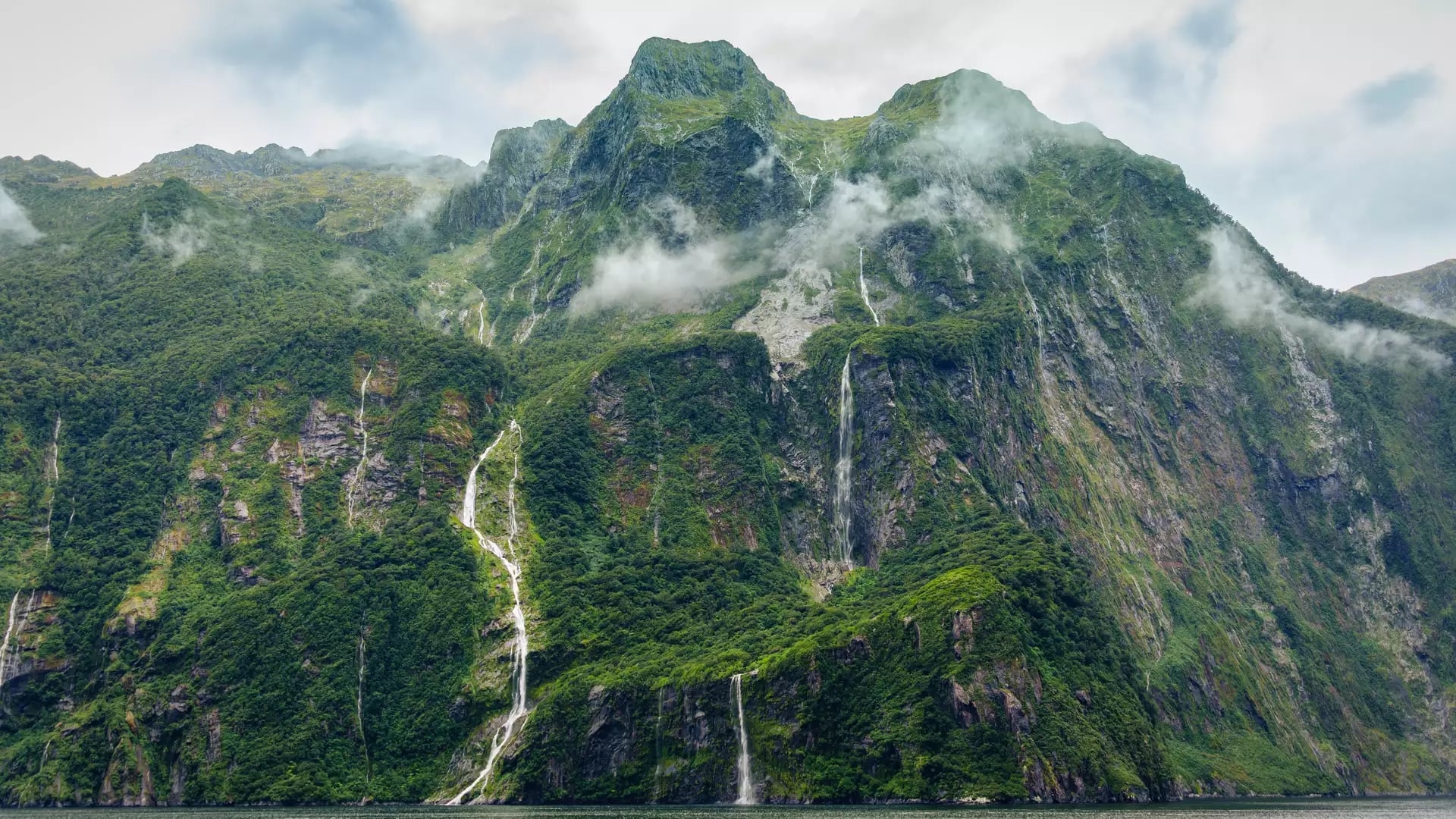 A towering green mountain cliff in Milford Sound, New Zealand, featuring multiple waterfalls cascading into the dark fiord water below, with wisps of low fog hugging the rainforest slopes under an overcast sky. Credit: Mumemories - Shutterstock