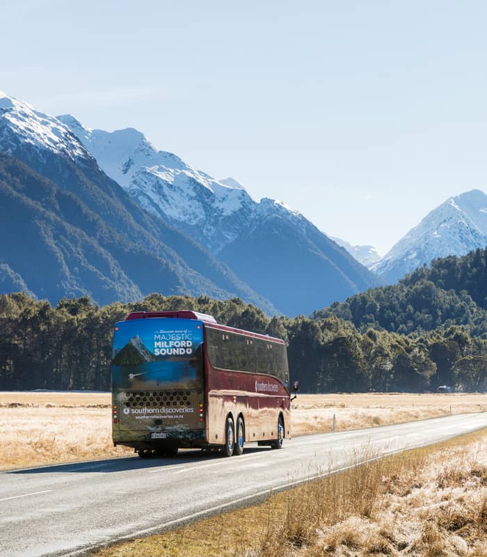 Coach driving on a road through Hollyford Valley with snow-capped mountains in the background.