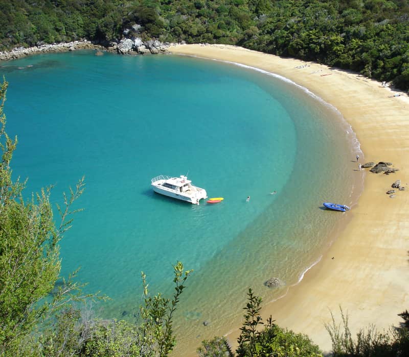 Turquoise bay with sandy beach, anchored boat, kayaks, and surrounding forest.