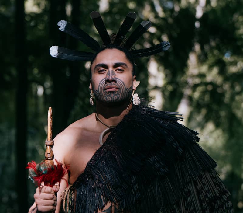 Maori warrior with tattoos and traditional attire holding a spear in a forest setting. Credit: Te Pa Tu