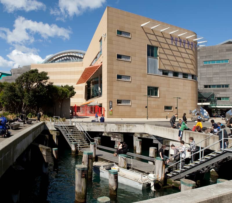People sitting and standing by the dock and benches outside the modern Te Papa Museum by the water. Credit: Museum of New Zealand Te Papa Tongarewa