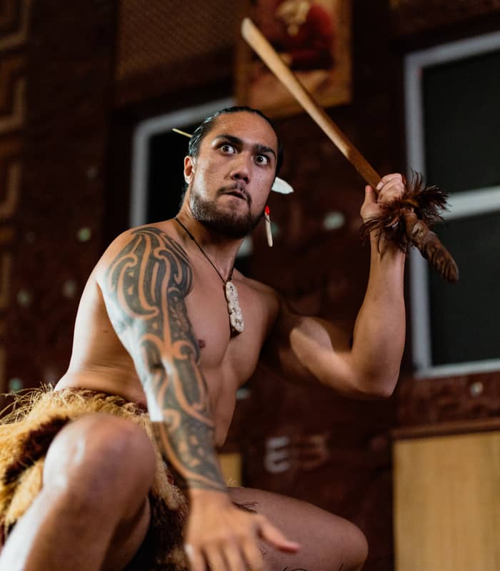 Maori man with tattoos holding spear performing traditional dance indoors. Credit: Te Puia