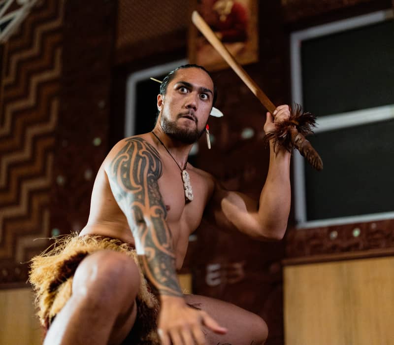 Maori man with tattoos holding spear performing traditional dance indoors. Credit: Te Puia
