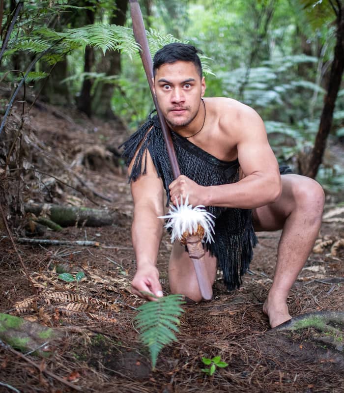 Maori man in traditional attire crouched in forest holding a spear with white feathers. Credit: Waitangi Treaty Grounds