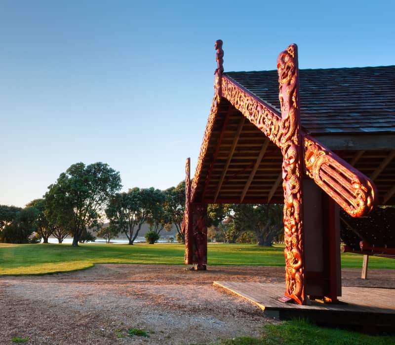 Sunset lighting up a carved Maori shelter near water and trees on a clear day. Credit: Waitangi Treaty Grounds