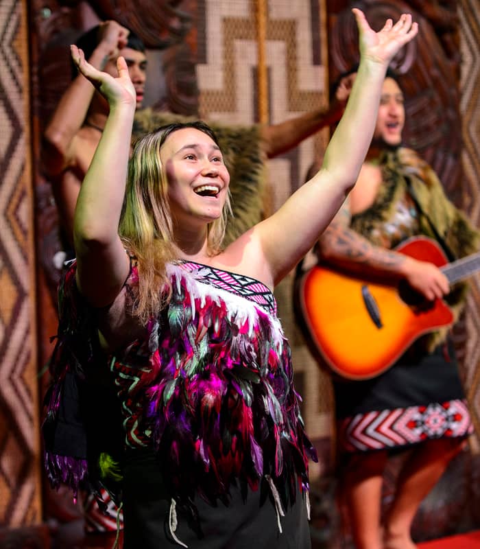 Smiling woman in traditional Māori feathered dress raising arms during cultural dance performance with guitar player in background. Credit: Waitangi Treaty Grounds