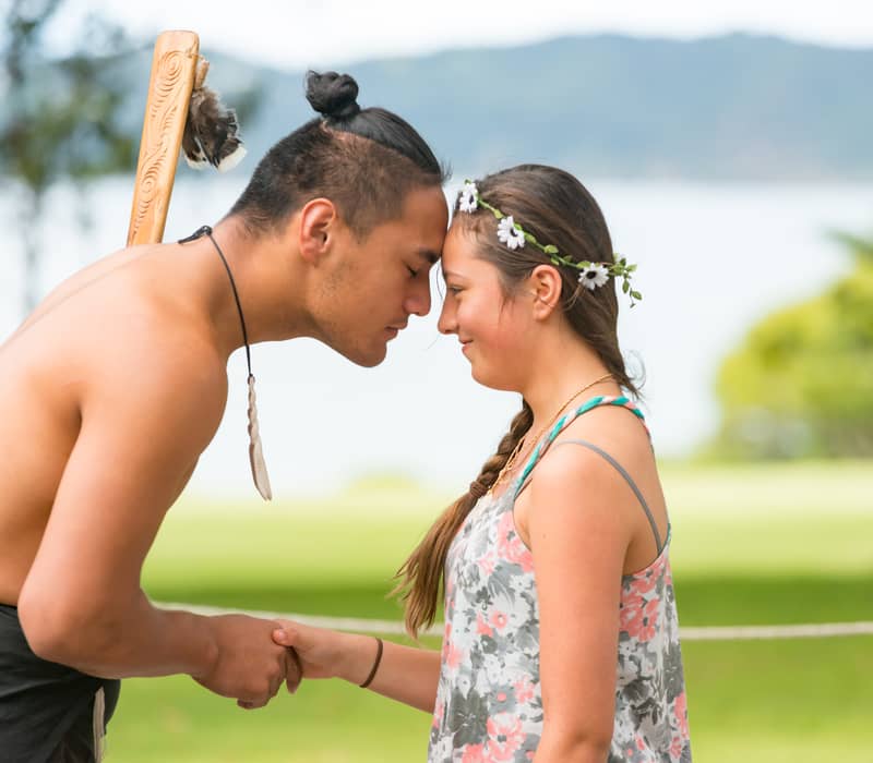 A young man and woman touching foreheads and holding hands in a traditional Maori greeting outside. Credit: Waitangi Treaty Grounds