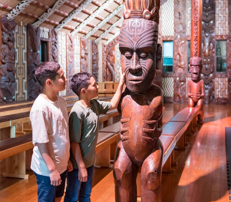 Two boys looking at and touching a large carved wooden Maori statue inside a decorated hall. Credit: Waitangi Treaty Grounds