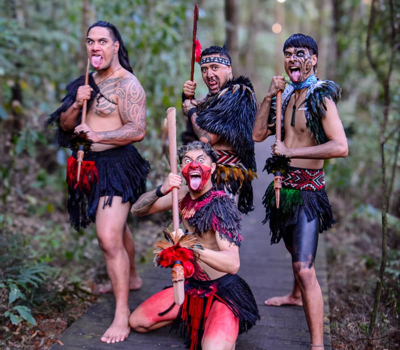 Four Maori men in traditional attire on a forest path making fierce expressions with spears. Credit: Waitangi Treaty Grounds