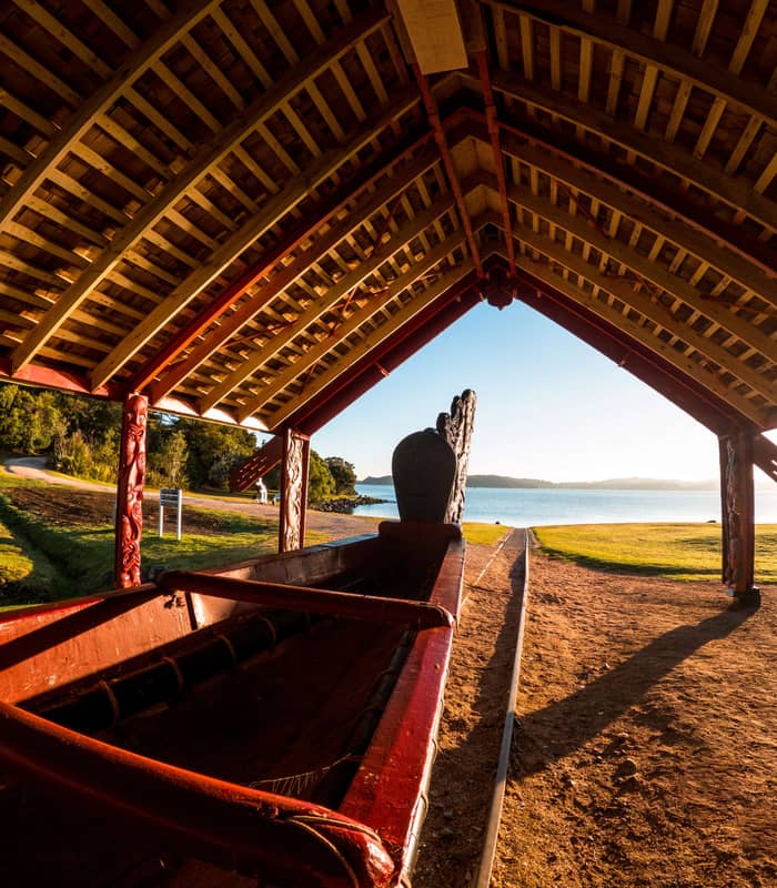 Traditional carved waka under a wooden shelter by a lake at sunset with grassy area and trees. Credit: Waitangi Treaty Grounds