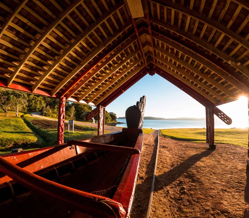 Traditional carved waka under a wooden shelter by a lake at sunset with grassy area and trees. Credit: Waitangi Treaty Grounds