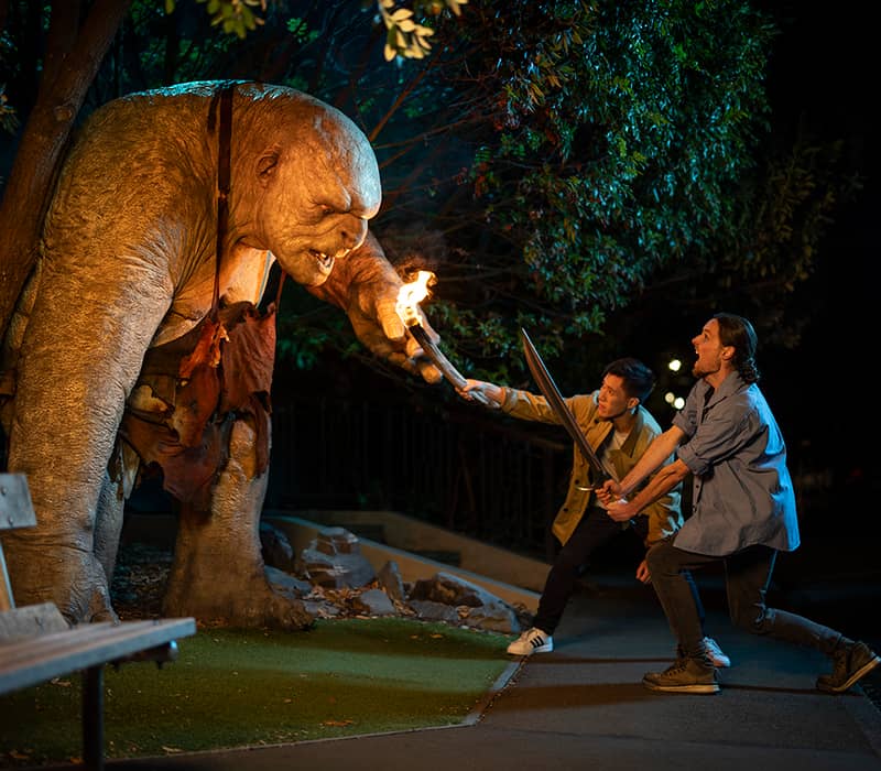Two men with swords face a giant troll statue holding a flaming torch at night outdoors.