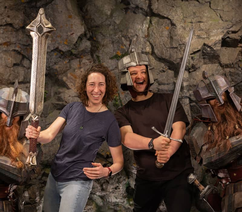 Two people smiling and holding swords while wearing metal helmets, posing with dwarves in armor against a rocky background. Credit: Weta Workshop