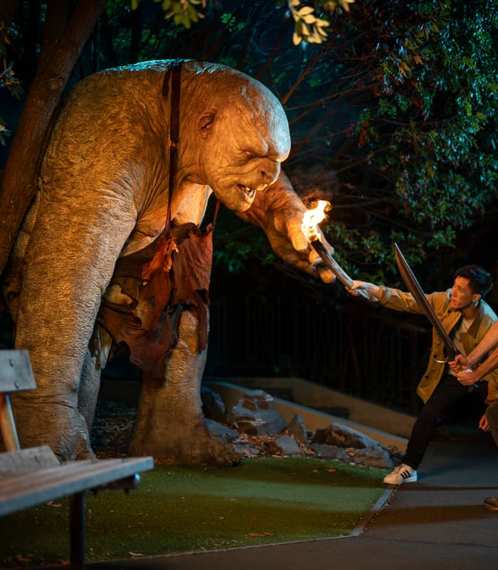Two men confronting a large troll sculpture holding a torch at night in a park setting. Credit: Weta Workshop