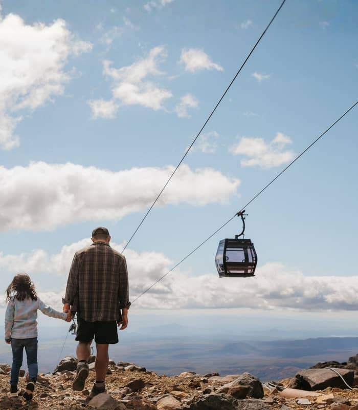 Adult and child holding hands hiking on rocky mountain trail near cable car under blue sky with clouds. Credit: Whakapapa Holdings 2024 Limited